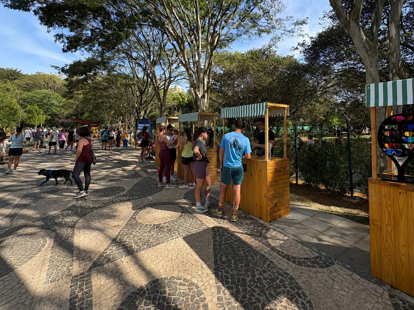 Público circulando entre estandes de madeira na alameda arborizada da Orla TotalPass